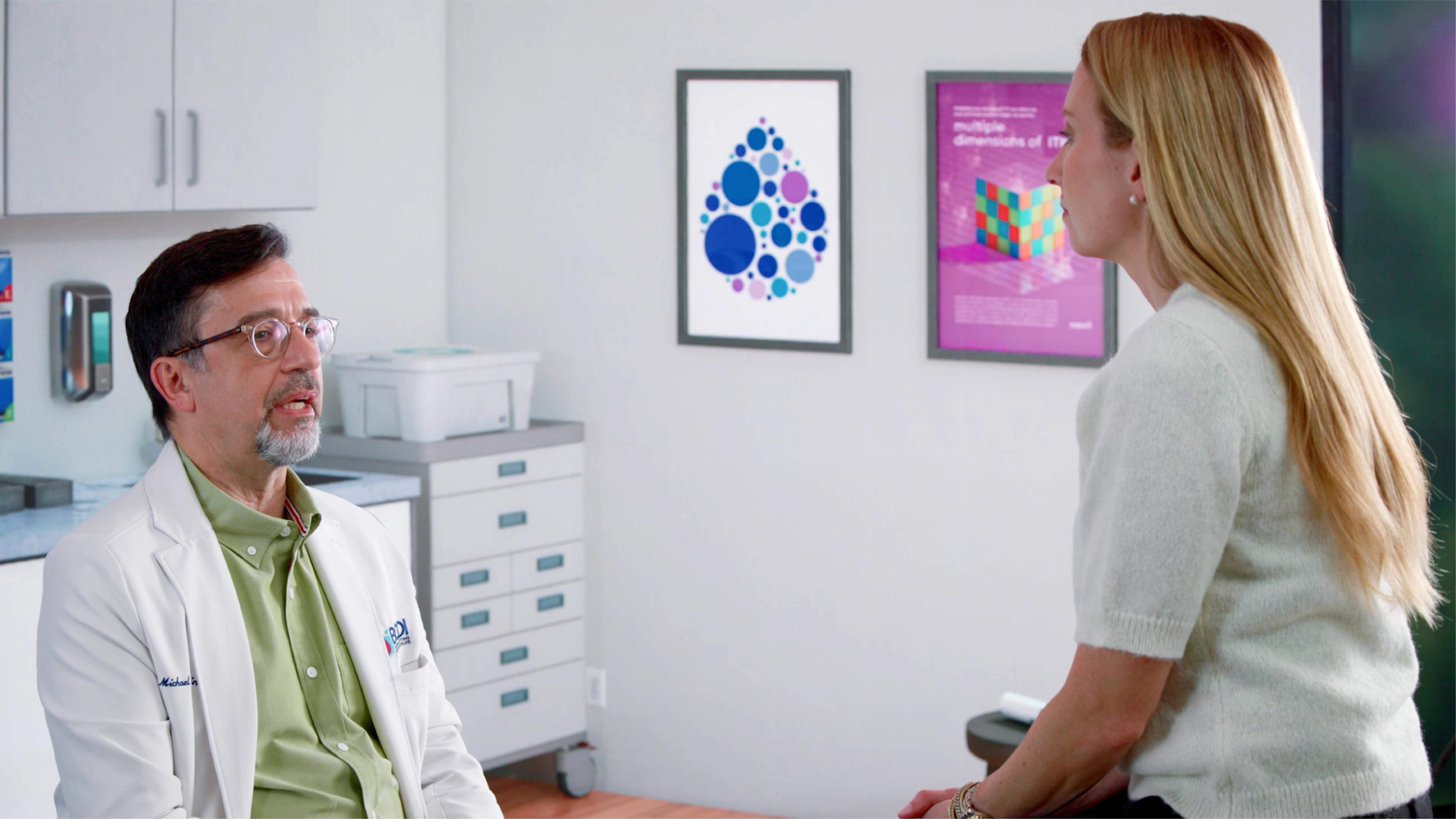 Holly, wearing a white sweater, sits in an exam room turned away from the camera and discusses her care plan with Dr. Tarantino, her provider, who is wearing glasses, a green shirt and white lab coat. 