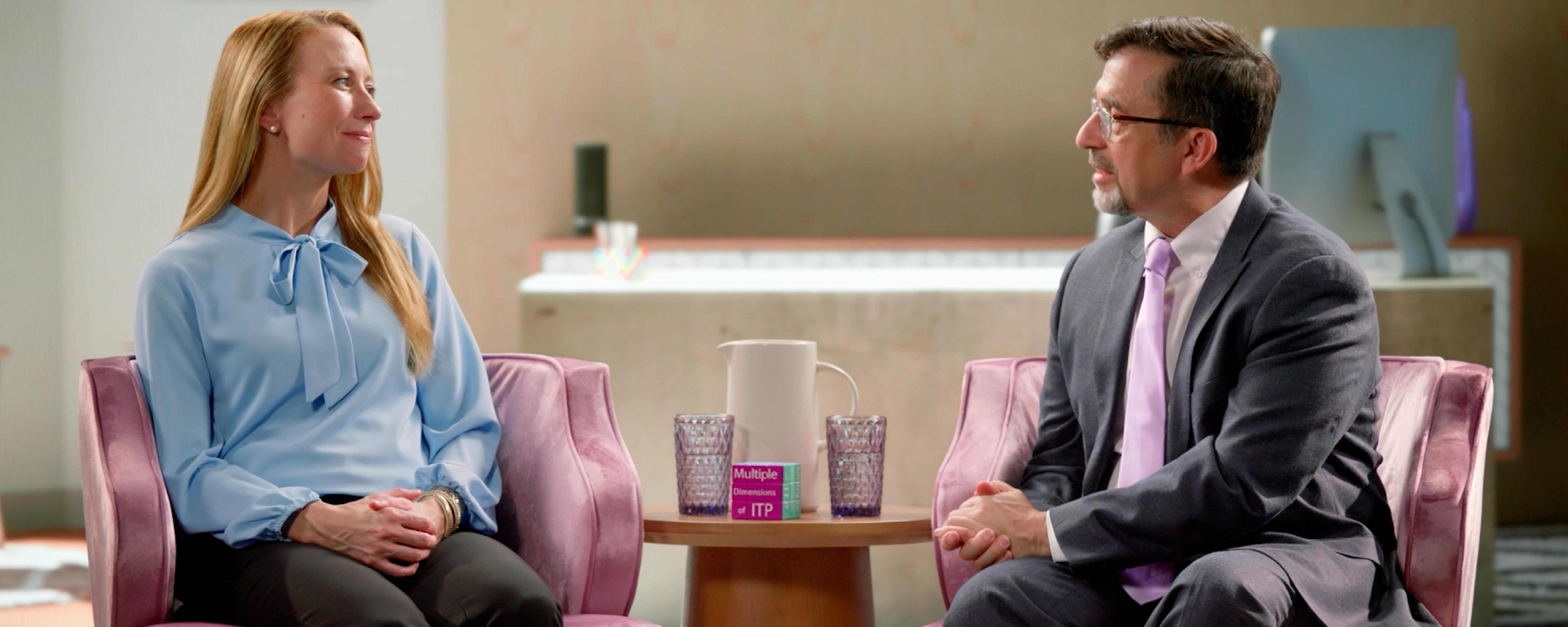 Holly, wearing a blue blouse, sits in an armchair in a living room setting with her provider, Dr. Michael Tarantino, wearing glasses, a grey suit, and a purple tie, during a conversation about ITP. 