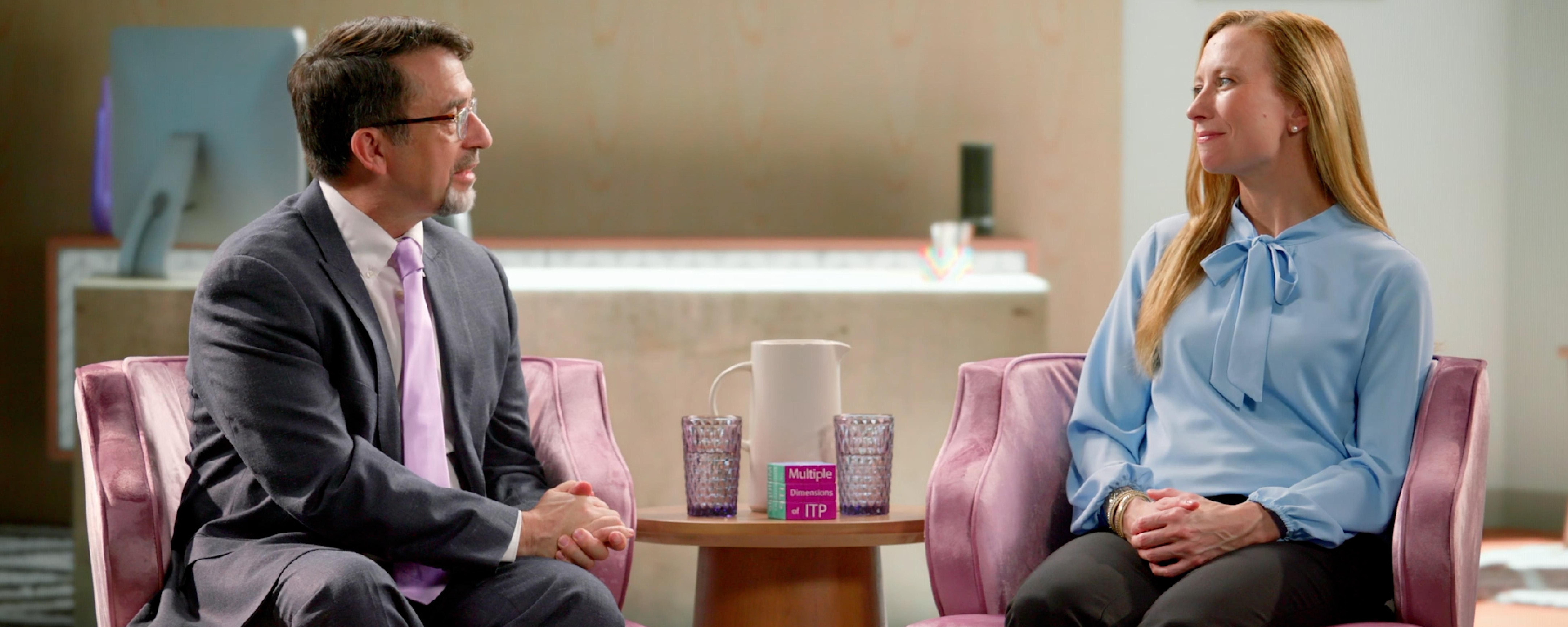Holly, wearing a blue blouse, sits in an armchair in a living room setting with her provider, Dr. Michael Tarantino, wearing glasses, a grey suit, and a purple tie, during a conversation about ITP.  