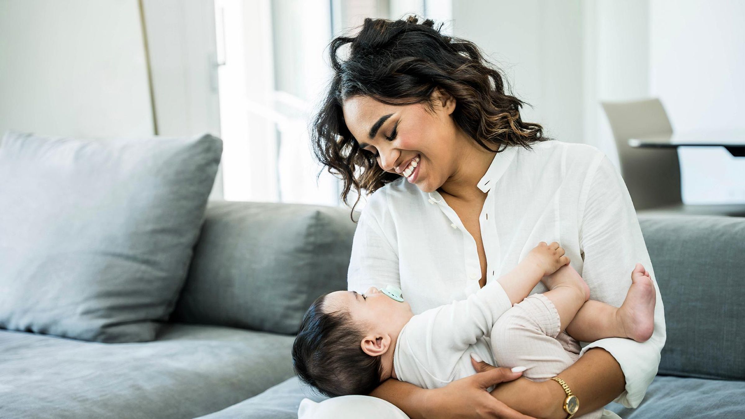 A mother cradling her infant on a couch at home, gazing down warmly as the baby looks up with a pacifier — a quiet moment from the early months of life, when RSV protection matters most.