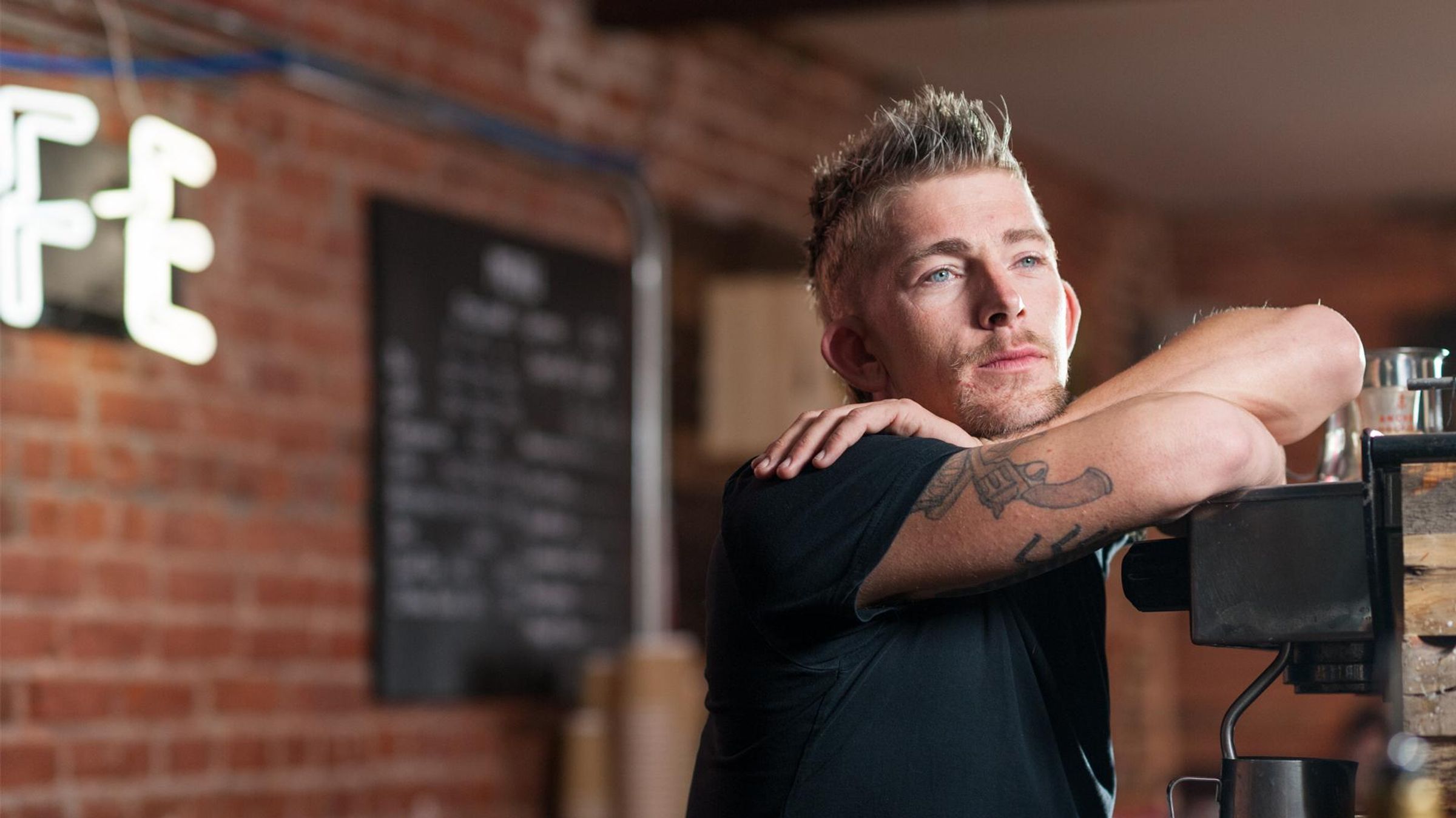 JJ, a man with visible tattoos on both arms, stands confidently with arms crossed while leaning against an exposed brick wall inside a restaurant. He wears a black shirt and has a calm, approachable expression. A menu is visible on the wall behind him. JJ lives with Acid Sphingomyelinase deficiency (ASMD), a rare disease, and is featured as part of Sanofi's commitment to the rare diseases community affecting over 350 million people worldwide