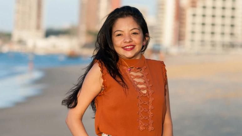 Portrait of Anna, a woman with long wavy black hair, standing on a beach and smiling at the camera. She wears an orange lace-detailed blouse, white floral skirt, and a pendant necklace. The background shows a blurred beach scene with sand, ocean, and buildings under a partly cloudy sky.