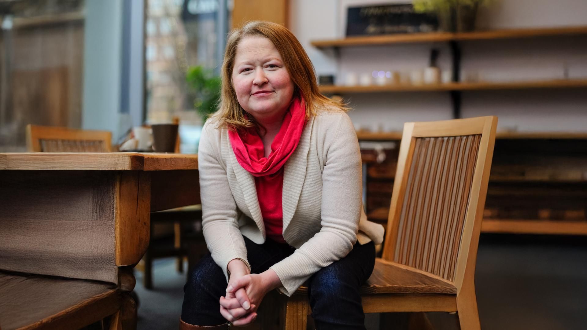 A woman smiling gently sitting on a wooden chair in a café setting wearing a red scarf.
