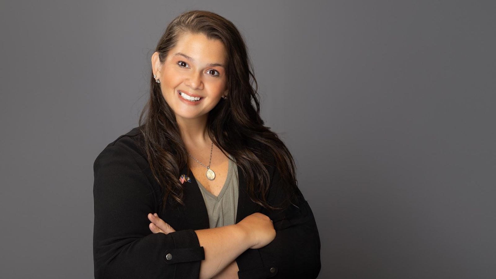 A woman posing against a dark gray backdrop smiling with her arm’s folded
