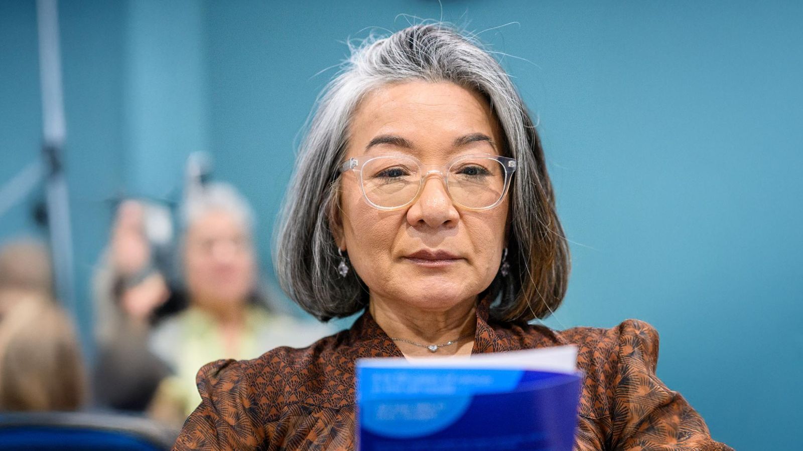 Senior woman with gray hair and glasses holding a blue folder, standing in front of blurred audience members at an indoor healthcare professional meeting.
