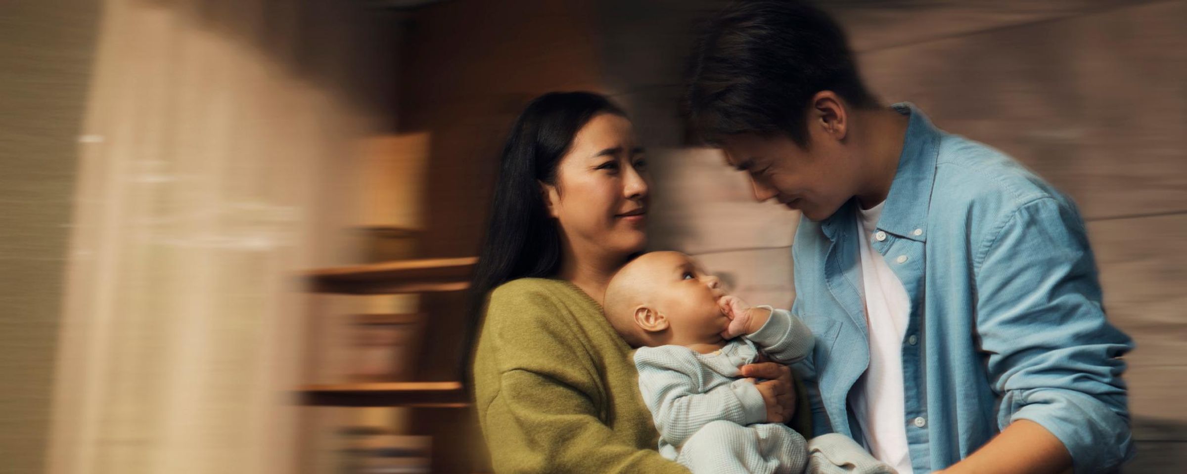 A content infant in a light blue knitted outfit rests on a soft blanket while a caregiver gently holds the baby's feet, illustrating the protective bond between caregiver and child. The warm, nurturing scene emphasizes infant care and health protection