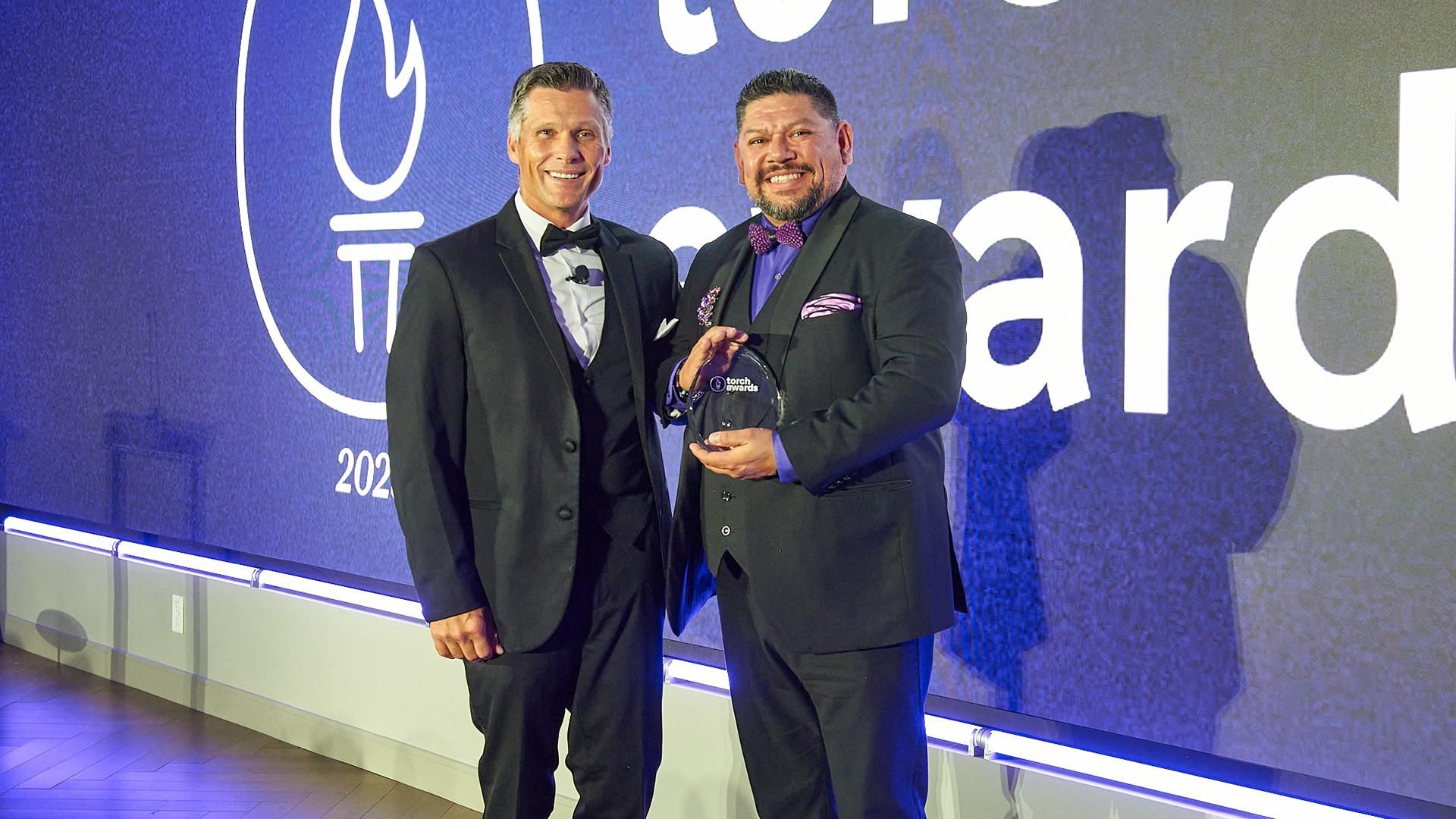 Two men in tuxedos at the Sanofi TORCH Awards stand in front of a screen smiling while one holds his award.