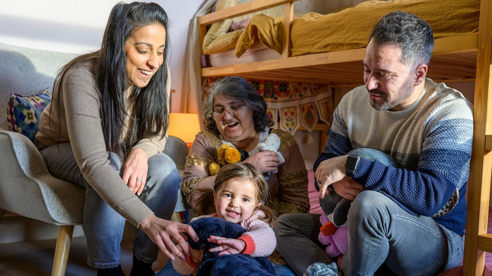 A multi-generational family of a toddler girl, mother, father and grandmother play in the toddler’s room.