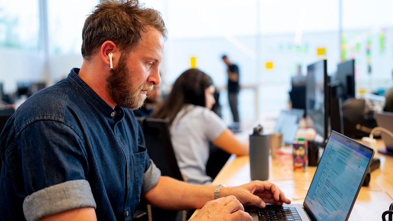 Sébastien Renard, with a blue jean shirt, blond hair and beard, working on his laptop wearing ear-pods in a shared workspace.