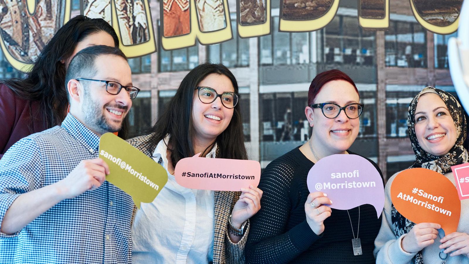 A group of Sanofi employees smiles while holding up signs that read “sanofi@Morristown” and “#SanofiAtMorristown”.