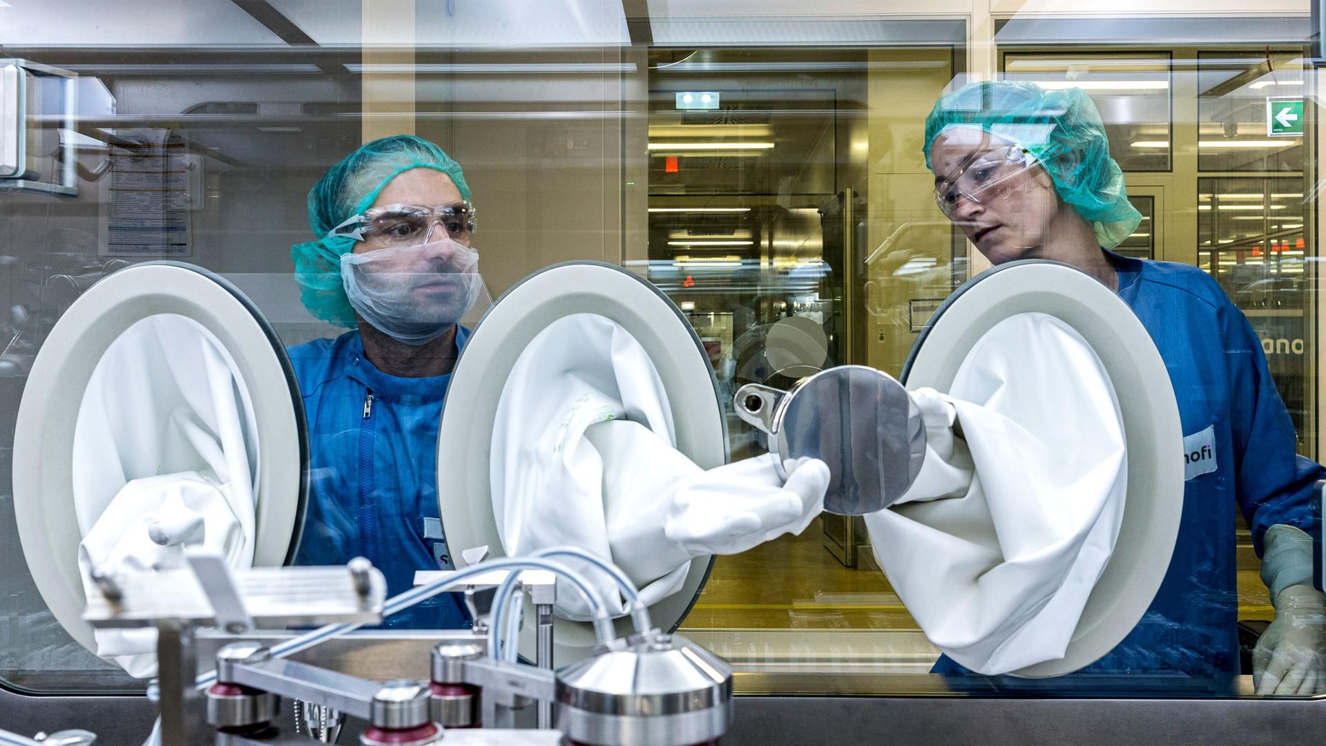 Emre Canaki and Stefanie Kutschmann pass a metal disk between them by way of gloveboxes inside a large steel machine.