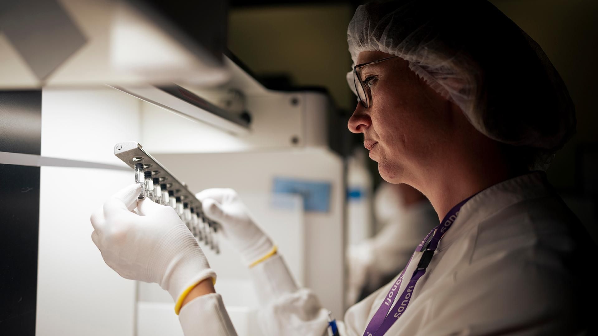 Nadège Bailleul inspects a set of 12 small clear vials on a steel rack next to a fume hood in a darkened room.