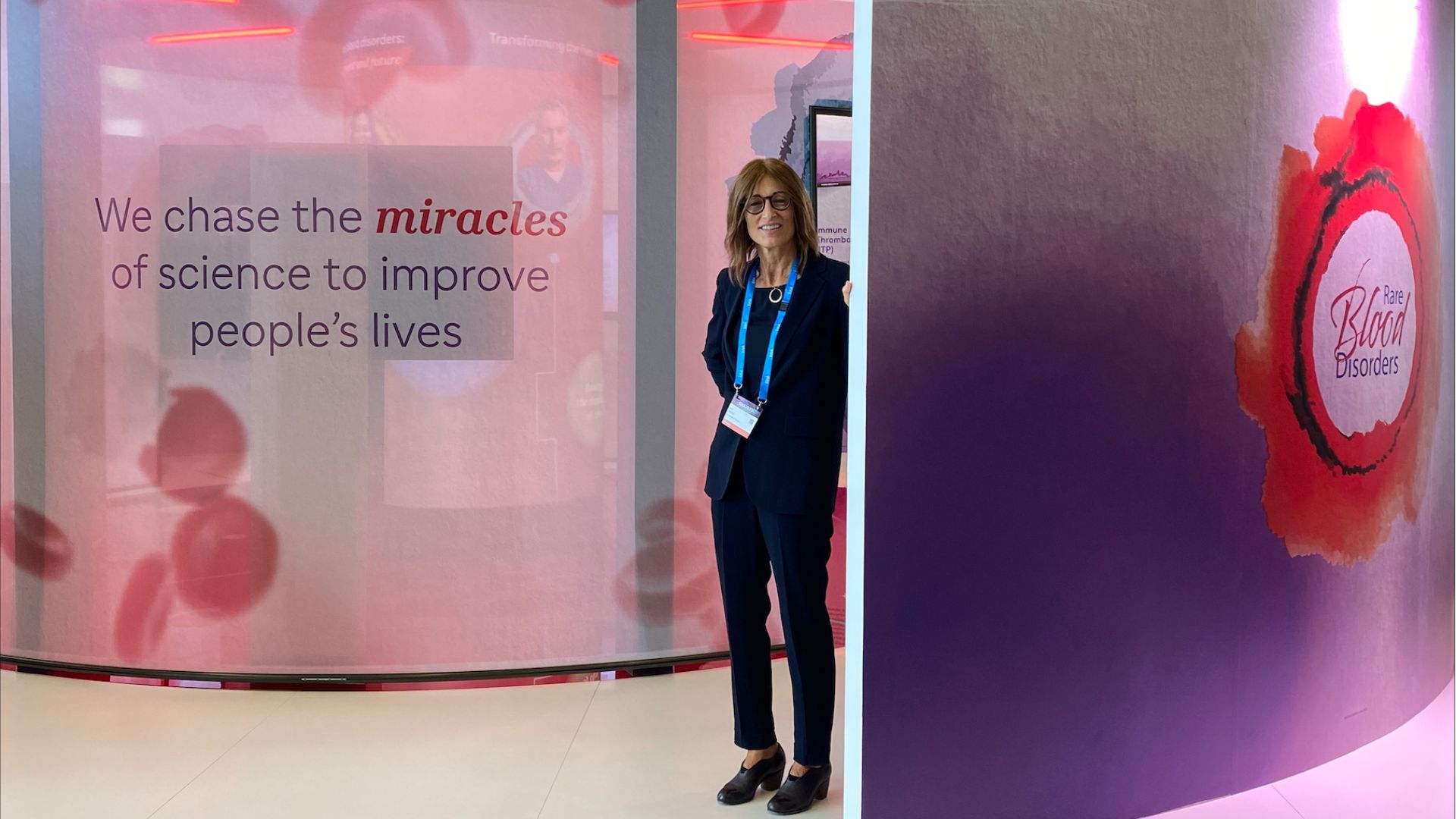 Dr. Karin Knobe, dressed in a suit with a lanyard, is standing in a medical conference booth that has images of red blood cells; on the left, there is text that says “We chase the miracles of science to improve people’s live”; on the right, there is text that says “Rare Blood Disorders.”