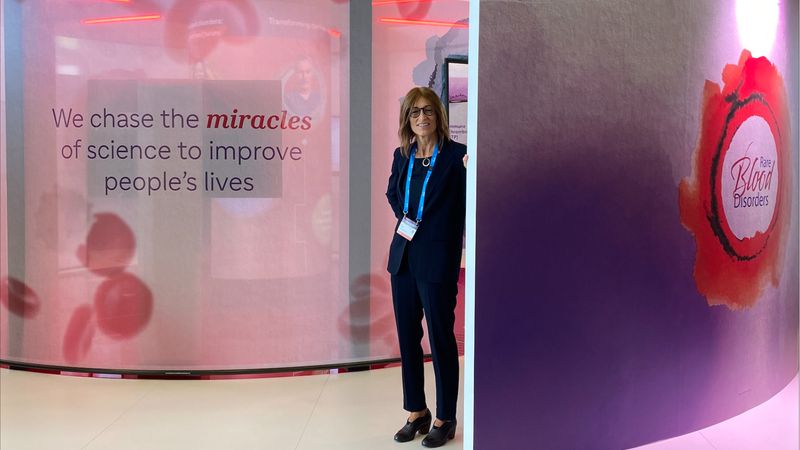 Dr. Karin Knobe, dressed in a suit with a lanyard, is standing in a medical conference booth that has images of red blood cells; on the left, there is text that says “We chase the miracles of science to improve people’s live”; on the right, there is text that says “Rare Blood Disorders.”