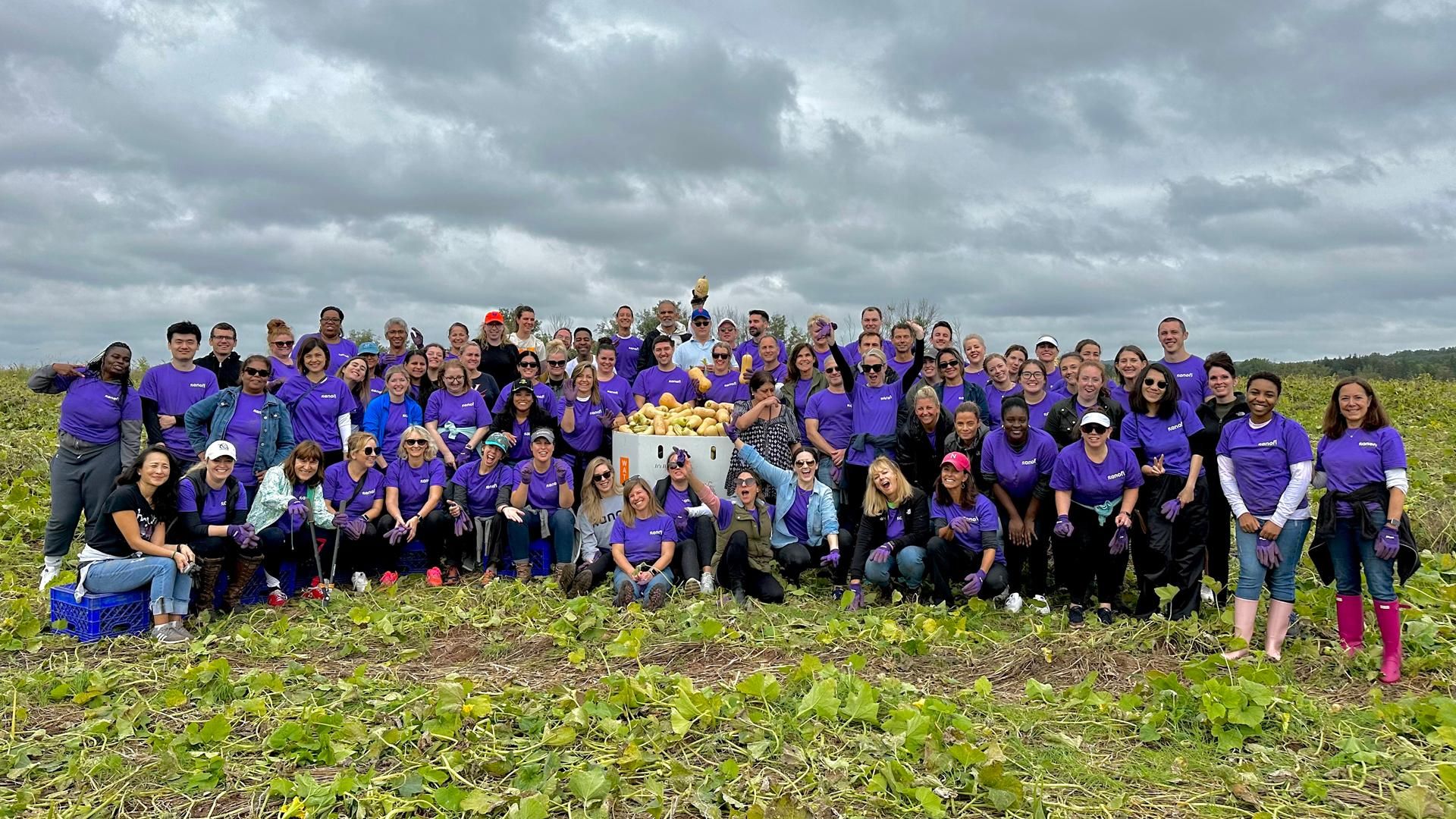 A large group of people all wearing purple shirts sit or stand in a field holding up squashes after a volunteering activity picking squash at a farm.