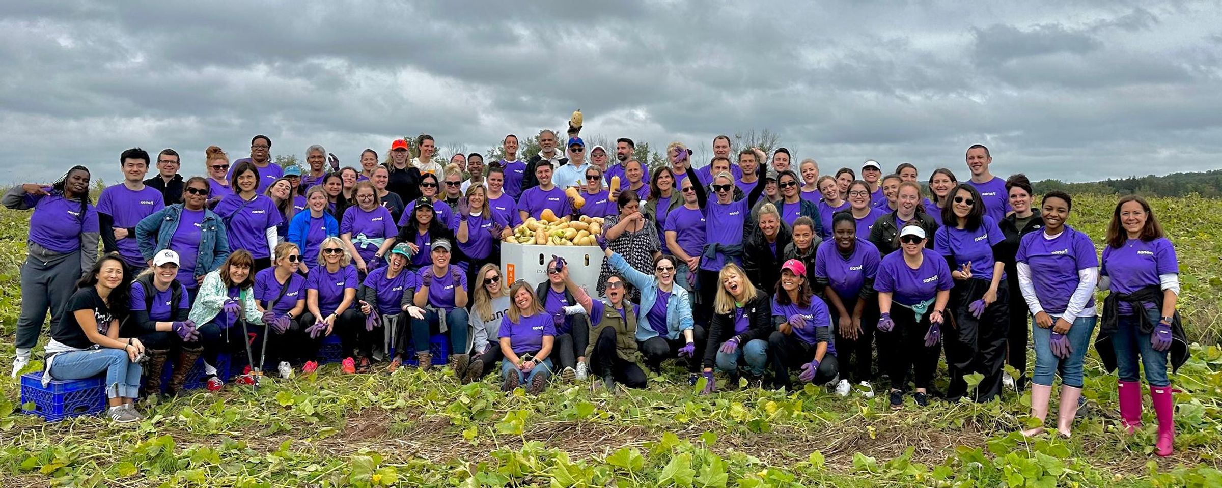 a large group of people in purple shirts standing in a field smiling after volunteering at a farm to pick squash.
