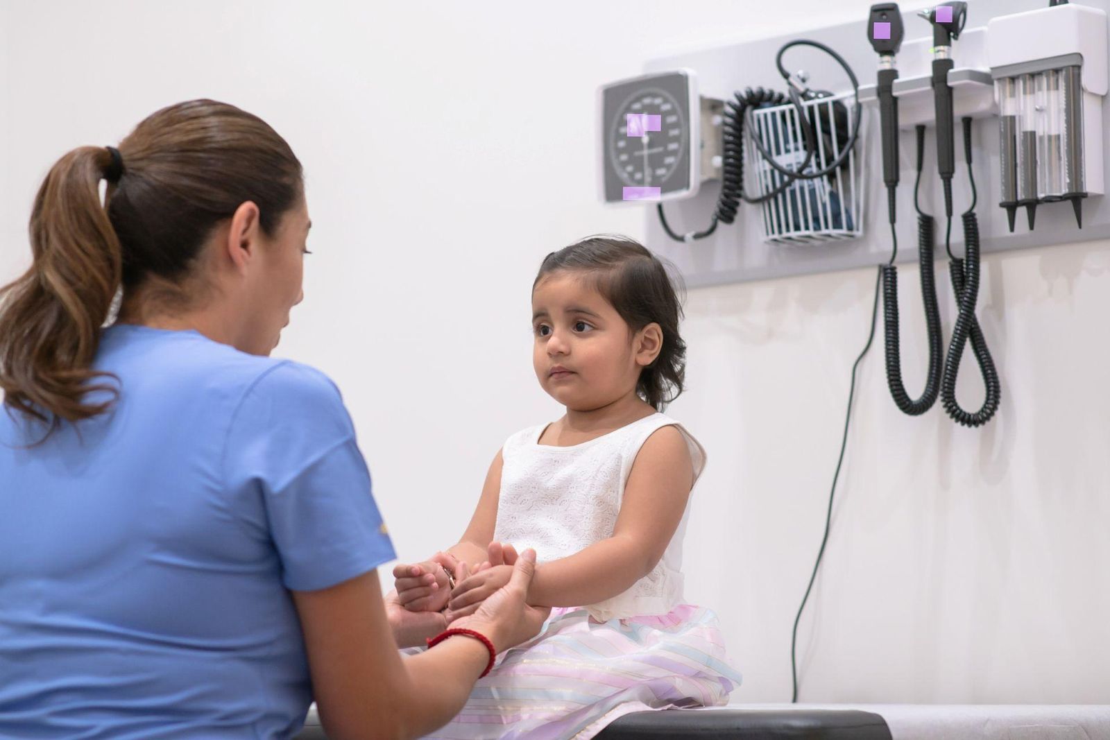 A healthcare provider speaks with a young child seated on an exam table in a clinical setting.