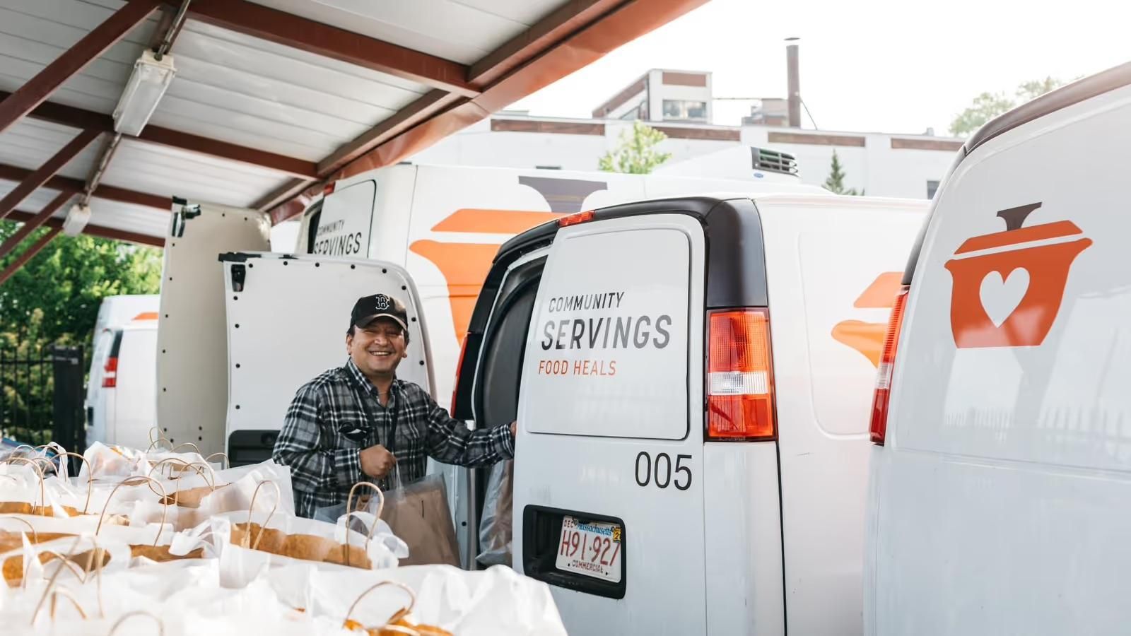 A Community Servings team member stands beside a branded delivery van with bags of prepared meals ready for delivery.