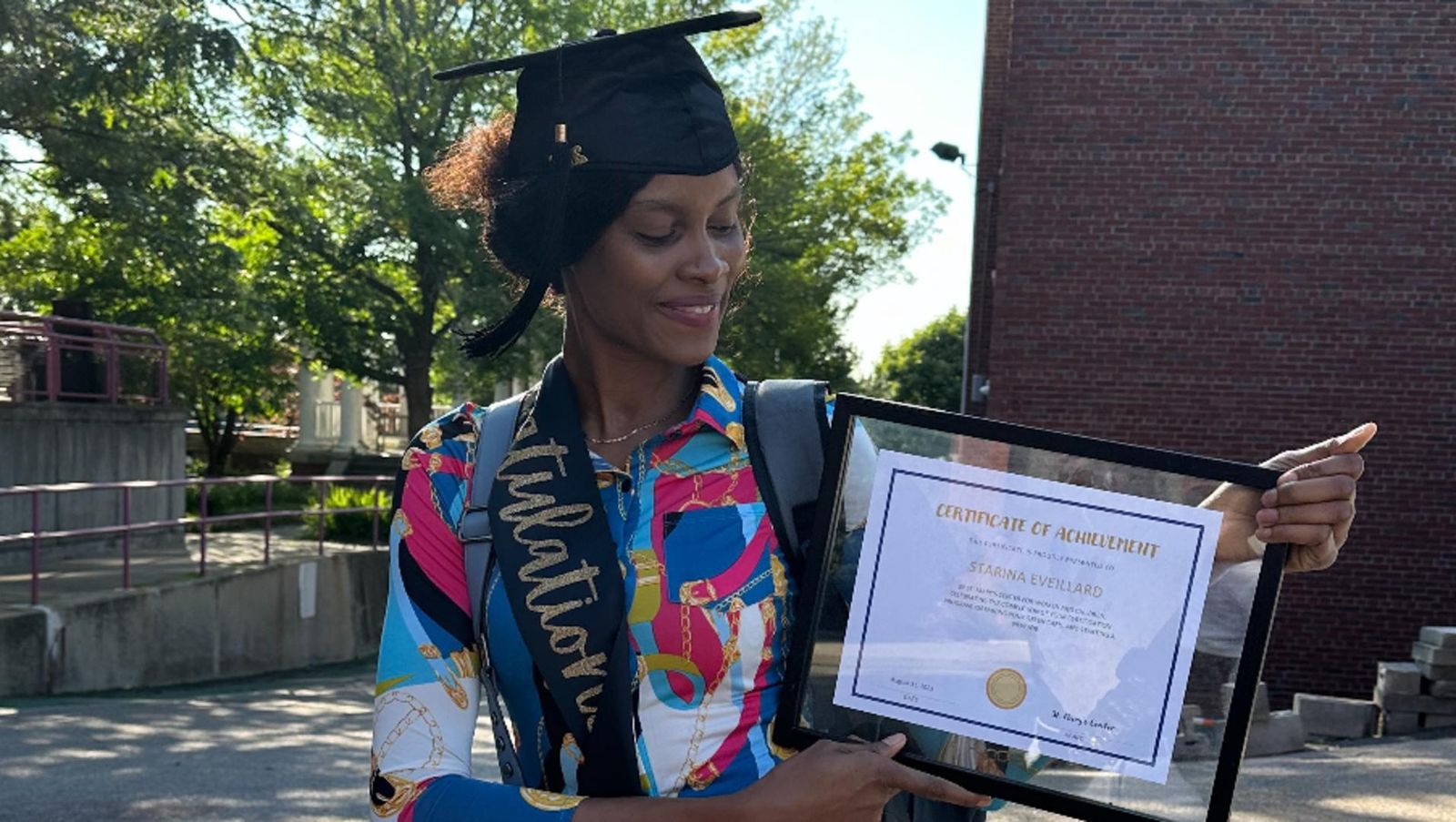 A woman wearing a graduation cap and sash holds a framed certificate of achievement outdoors.