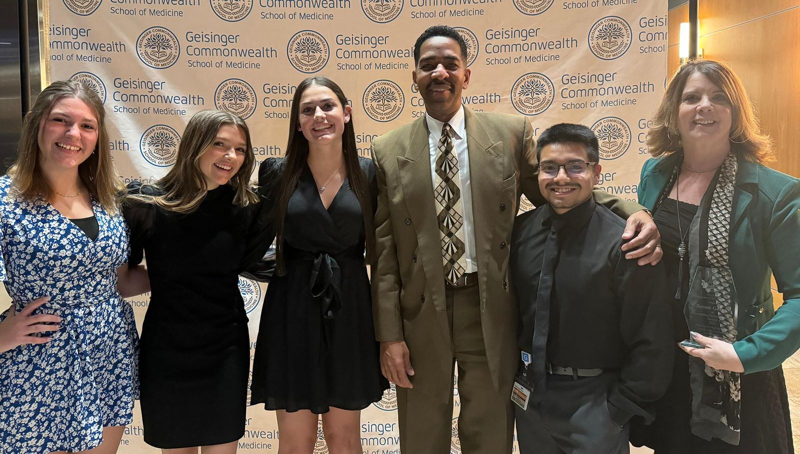 Group photo of six people standing in front of a Geisinger Commonwealth School of Medicine banner. From left to right: a woman in a blue floral dress, a woman in a black dress, another woman in a black dress with hands on hips, Phillip St. James in a dark suit and tie in the center, a woman in a black shirt and green blazer, and a woman in a black dress and green blazer. The white banner behind them displays the school's name and logo multiple times, featuring a shield with a tree and book symbol.