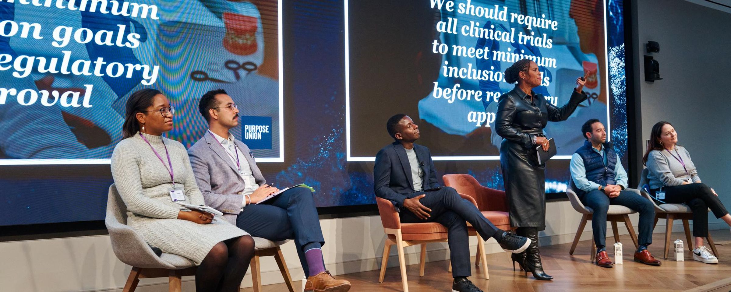 Sanofi employees participate in "The Art of Dialogue" professional development session led by facilitators Lewis Iwu and Tanisha Sullivan. Female presenter in black jacket leads interactive workshop on navigating disagreement and persuasion techniques. Five participants seated on stage in modern Sanofi conference room with presentation screen displaying dialogue and communication strategies. Corporate training focused on effective workplace communication, conflict resolution, and persuasive dialogue skills for pharmaceutical industry professionals.