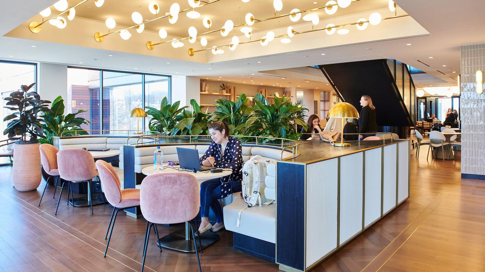 Interior view of Sanofi's modern Morristown, New Jersey office café and collaborative workspace. Employees work on laptops at communal tables surrounded by large potted plants. The space includes a sleek counter with geometric wood paneling, natural light from large windows, and a multi-level design with visible staircase. This innovative workplace environment reflects Sanofi's commitment to employee wellbeing, collaboration, and modern pharmaceutical industry workspace design in the United States.