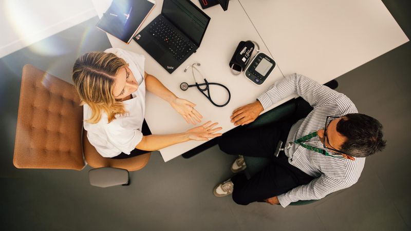 Overhead view of a desk with a healthcare professional and a patient in conversation, stethoscope and laptop visible.