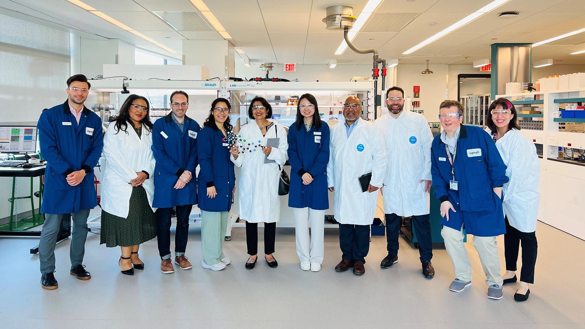 Nine diverse Sanofi professionals wearing lab coats—mostly blue with some white—standing together and smiling in a modern laboratory. The research team members wear name tags and lanyards, positioned in front of laboratory equipment and shelving, conveying collaboration and inclusive teamwork in pharmaceutical research.