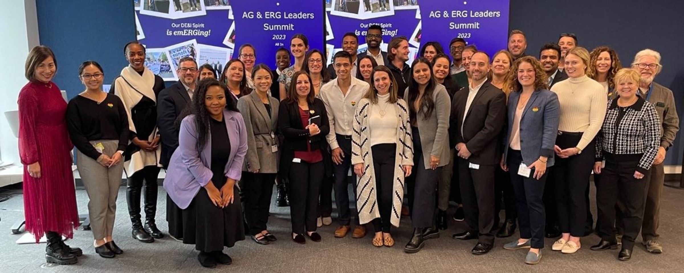 A group photo of approximately 20 Sanofi employees gathered at the AG & ERG Leaders Summit 2023. The diverse group of professionals, varying in gender, race, and age, stands together smiling in business attire. Behind them is a large banner reading "Our DEI! Spirit is emERGing!" and "AG & ERG Leaders Summit 2023." The indoor setting and professional atmosphere reflect a collaborative event celebrating the company's and inclusion initiatives through its Affinity Groups and Employee Business Resource Groups.
