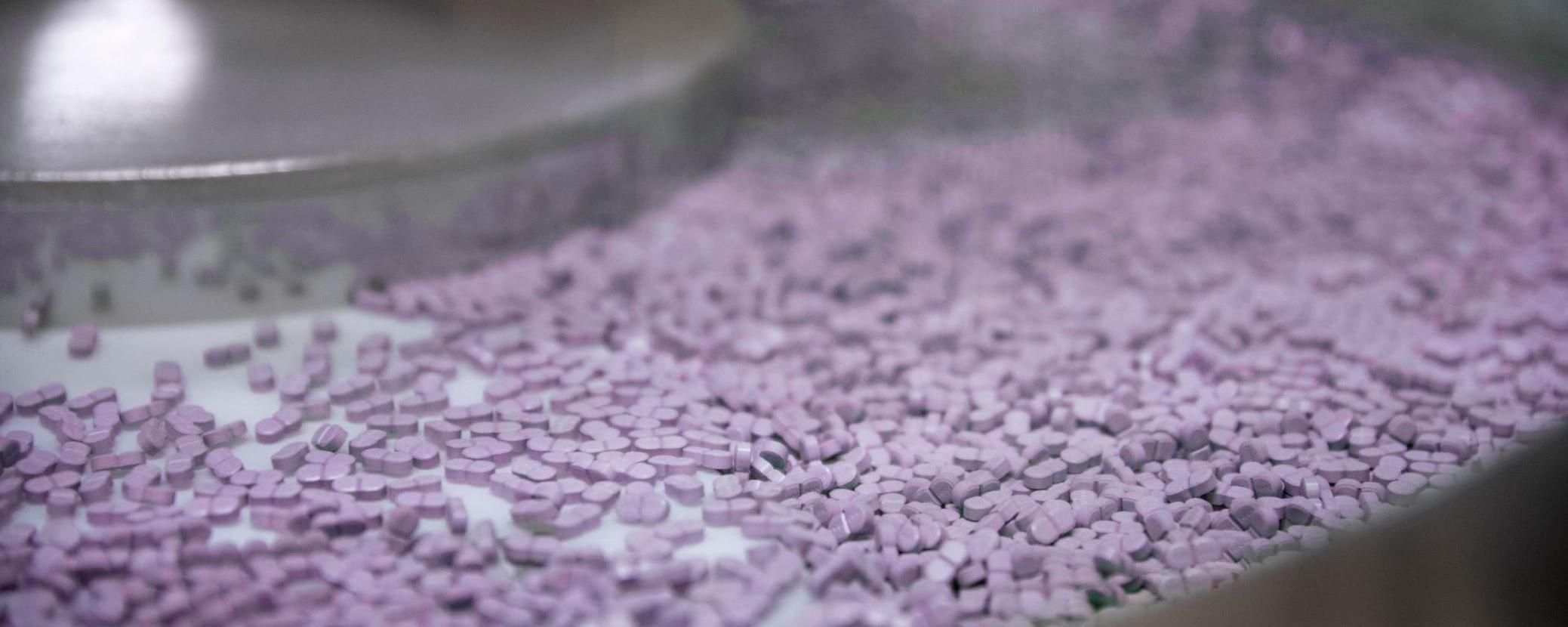 Conveyor belt filled with numerous small, purple tablets in a manufacturing setting