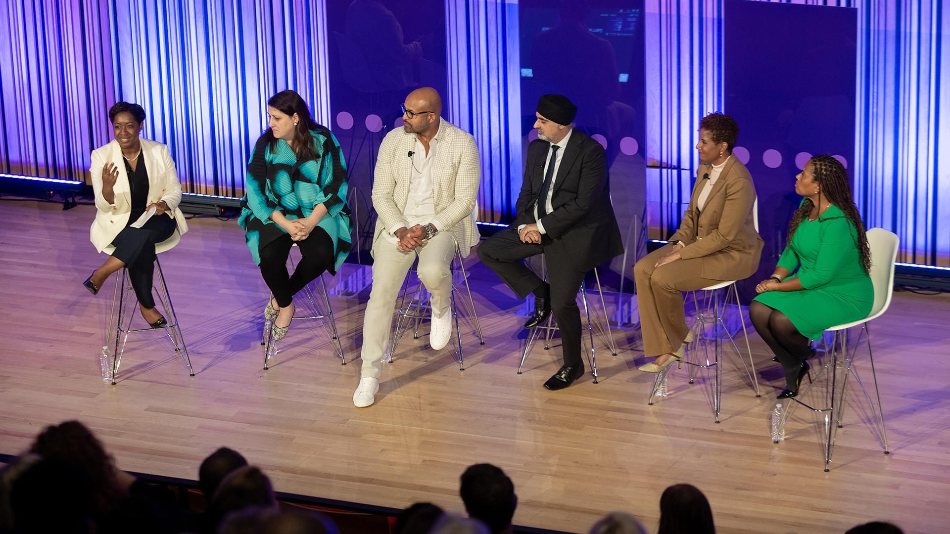 A group of 4 women and 2 men sit on the stage speaking to a crowd.
