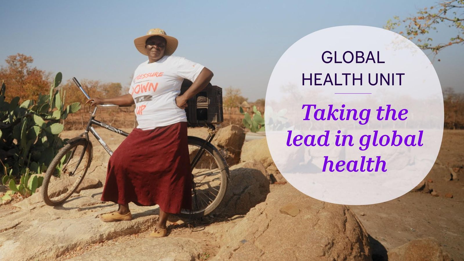 A black woman in a straw hat, white t-shirt and red skirt stands on rocky ground while holding her bike upright behind her.