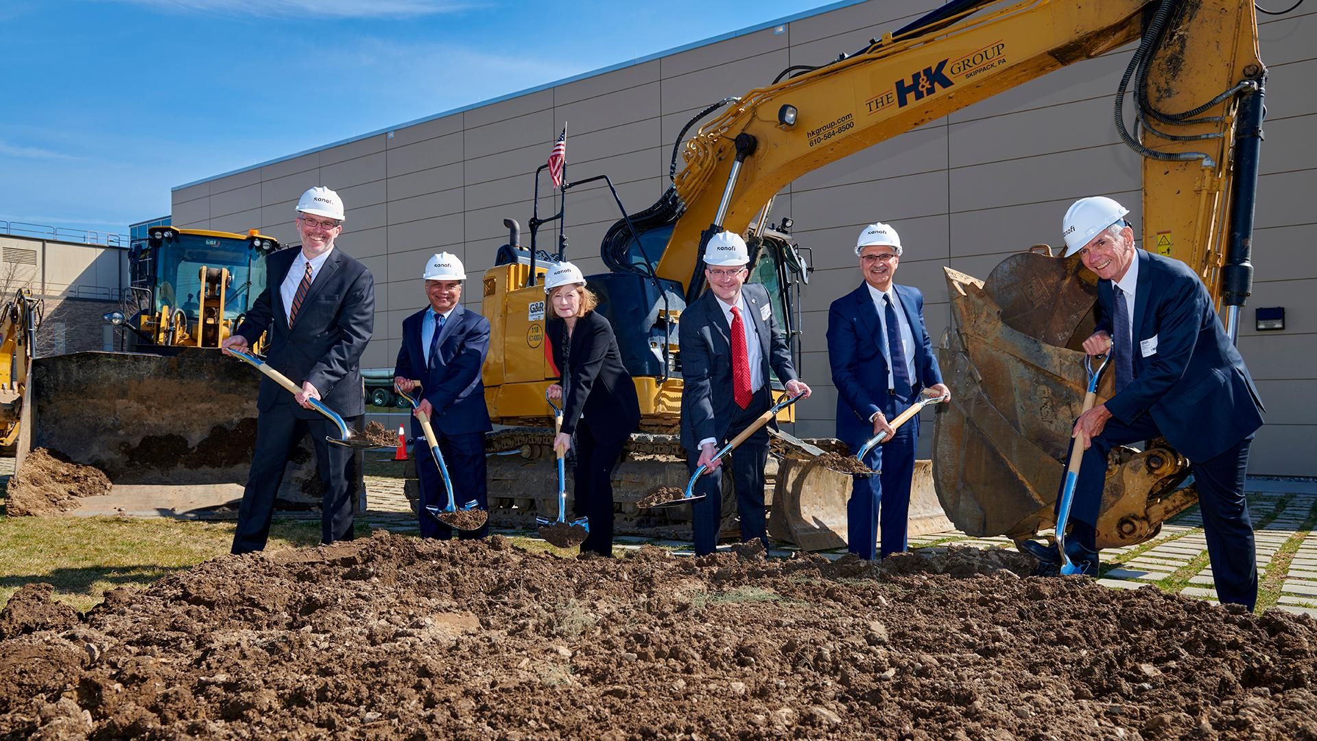 Six Sanofi executives and officials in business attire and white hard hats hold ceremonial shovels during the groundbreaking ceremony for Sanofi's new sustainable pandemic flu vaccine production facility in Swiftwater. An HK Group excavator with an American flag stands ready behind the group, with the facility building visible in the background under a clear blue sky.