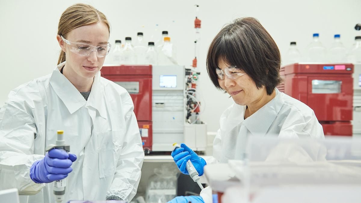 Two scientists in white lab coats, safety glasses, and blue nitrile gloves work side by side at a laboratory bench using pipettes. There is red scientific equipment and rows of large clear bottles visible in the background.