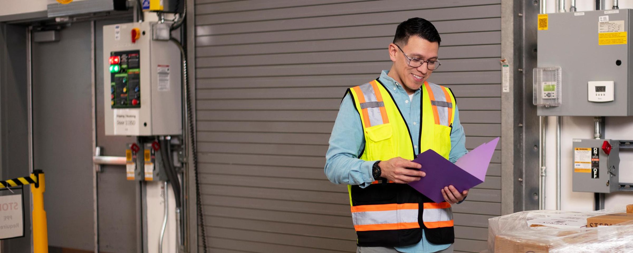 A worker in a high-visibility safety vest stands in a warehouse beside shrink-wrapped pallets of boxes, reviewing a purple clipboard in front of a large roll-up loading door and industrial control panels.