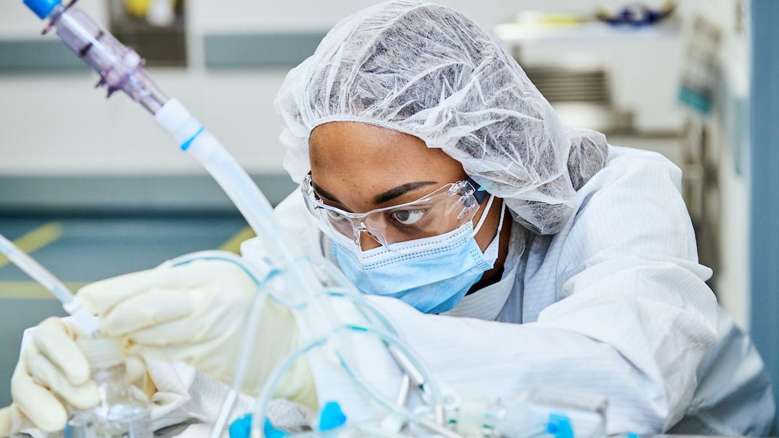 A laboratory worker wearing a hair cover, safety glasses, mask, and gloves closely adjusts equipment in a manufacturing lab.