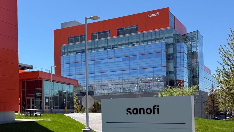 A modern multi-story Sanofi facility with a contemporary red and glass façade displaying the Sanofi name in white lettering. The building is surrounded by neatly trimmed grass, trees, and a picnic area, with a clear blue sky in the background.